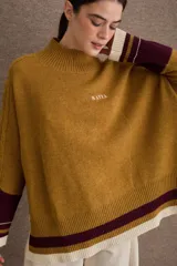 Buzo tejido de silueta amplia con cuello alto, mangas largas y puños en contraste. Presenta un diseño con franjas horizontales en el ruedo y detalles de tejido trenzado en las mangas.