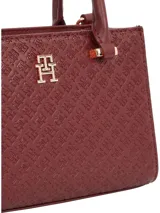 Cartera tote Tommy Hilfiger color borgoña con diseño integral del monograma TH en bajo relieve. Tiene doble asa corta, correa extraíble y ajustable, cierre de cremallera y herrajes dorados.