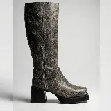 Bota de caña alta confeccionada en cuero vacuno, con diseño de punta cuadrada y plataforma. Presenta un taco cuadrado de madera, cierre lateral interno y una tira decorativa con herraje metálico circular en el tobillo.