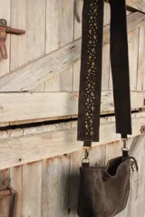 Bandolera confeccionada en cuero de gamuza marrón, con cierre superior. Presenta un llavero colgante con forma de estrella revestido en tachas metálicas y tiras flecadas en tono dorado.