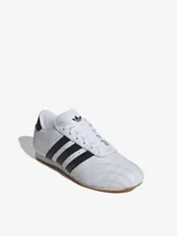 Zapatillas Adidas de cuero blanco con las tres tiras laterales en negro y suela de goma color caramelo.