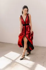 Vestido largo de seda color bronce con acabado brillante, escote profundo en V y espalda abierta. Presenta detalles en terciopelo negro que enmarcan el escote, recorren el frente y definen la cintura con una faja ajustable. Posee breteles anchos y una abertura frontal en la falda.