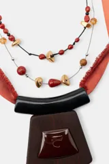 Pack de dos collares de diseño artesanal. El primero presenta una cinta de satén color terracota con flecos, adornada con cuentas negras y un colgante central de madera oscura con una pieza de resina incrustada. El segundo es un collar doble compuesto por una cadena metálica fina y un cordón negro, decorado con cuentas de piedra natural en tonos rojos, marrones y piezas metálicas doradas.
