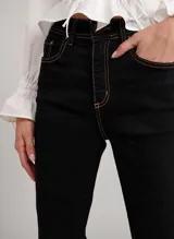 Pantalón de jean bootcut color negro con costuras en color naranja, tiro alto, pretina con trabillas, cierre frontal con cremallera y botón, y cinco bolsillos.