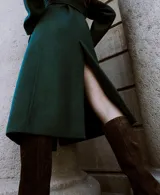 Abrigo largo de paño en color verde oscuro, con cuello alto, cierre frontal oculto y cinturón ajustable del mismo tejido. Presenta un diseño estructurado con bolsillos laterales.