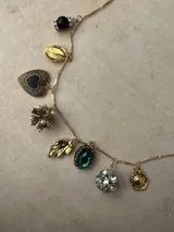 Collar de cadena fina en tono dorado con múltiples dijes colgantes, incluyendo una cuenta negra con cristales, hojas doradas, un corazón negro con borde texturizado, una flor dorada con perlas, un dije de piedra verde esmeralda, una esfera de cristales y un dije circular texturizado.