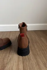 Botin de cuero vacuno color marrón, con suela track negra de goma y detalle de tirador en color rojo en la parte trasera.