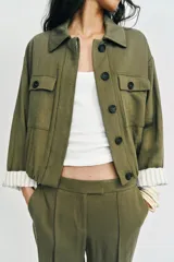 Chaqueta corta de color verde militar, con cuello solapa, bolsillos delanteros con solapa y cierre frontal de botones ocultos. Presenta mangas amplias con vuelta en los puños que muestran un forro a rayas.