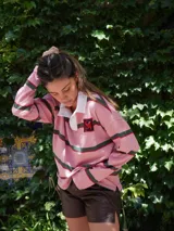 Buzo tipo rugby de algodón con diseño de franjas horizontales en tonos beige, negro y fucsia. Presenta cuello polo en contraste y cierre frontal parcial.