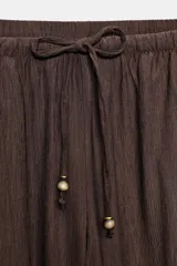 Pantalón negro de tiro alto con cintura elástica ajustable mediante cordones con terminales dorados. Presenta pernera ancha y bolsillos laterales. La tela tiene una textura sutilmente arrugada o plisada.