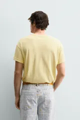 Camiseta de corte relajado color amarillo con efecto lavado. Presenta cuello redondo, mangas cortas con dobladillo y un estampado frontal con texto y un parche decorativo.