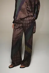 Pantalón de tiro alto confeccionado en seda, con cintura ajustable mediante cordones. Presenta un diseño de patchwork con diversos estampados geométricos y paisley en tonos tierra, verde y violeta.