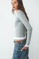 Camiseta de manga larga con diseño de doble capa, confeccionada en jersey suave y elástico. Presenta un calce ajustado, cuello redondo y un estilo minimalista que combina una capa superior en color verde grisáceo con una capa inferior en color blanco.