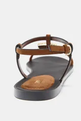 Sandalia plana con tiras de color marrón y un gran adorno metálico dorado circular en la parte frontal. Cuenta con cierre de hebilla ajustable al tobillo y suela plana de color negro.