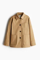 Chaqueta de algodón grueso color camel, con cuello camisero, cierre frontal de botones oscuros y cuatro bolsillos parche frontales (dos superiores y dos inferiores). Presenta hombros ligeramente caídos y puños con abertura de botón.