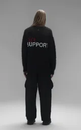Sweater de punto negro con textura suave, cuello redondo y diseño oversize. Presenta un bordado frontal con la frase 'art SUPPORT' en letras rojas y blancas, y un pequeño detalle de tijeras bordado en la nuca.