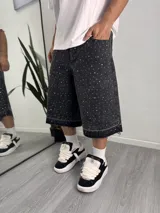 Bermuda baggy de denim negro con lavado ácido, corte holgado y diseño largo por debajo de las rodillas. Cuenta con apliques de strass distribuidos por toda la prenda y dobladillo deshilachado.