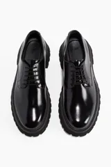 Zapato tipo Derby de cuero negro charol, con cordones y puntera redonda. Presenta una suela track gruesa y geométrica de goma negra.