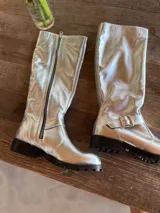 Bota alta plateada de cuero sintético con suela track negra, cierre lateral con cremallera y detalle de hebilla ajustable en el tobillo.