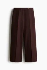 Pantalón sastrero de tiro alto color marrón, modelo tobillero de piernas amplias, con cierre frontal, pretina prolongada con botón y pliegues en la parte delantera y trasera.