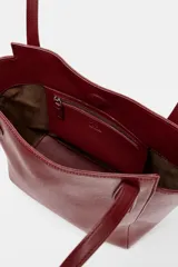 Bolso tipo shopper mini de color rojo oscuro o borgoña, con doble asa de hombro y pespuntes visibles. Cierre principal con botón imantado y bolsillo interior con cremallera.