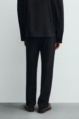Pantalón negro de corte recto (regular fit) confeccionado en tejido compacto. Presenta pliegues frontales marcados, cierre frontal con botón y cremallera, y bolsillos de vivo en la parte trasera.