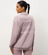 Buzo deportivo de corte oversized color lila, con cuello polo y cierre frontal con botones a presión. Presenta mangas largas con puños ajustados y una estampa bordada en el pecho. Confeccionado en una mezcla de poliéster, modal y elastano.