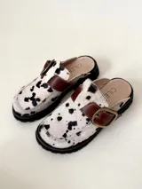 Zueco tipo mule destalonado con pelo estampado vaca (blanco y negro) y tira de cuero marrón con hebilla metálica. Posee suela negra de plataforma gruesa.