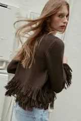 Chaqueta de punto color marrón oscuro, con cuello redondo y manga larga. Presenta flecos decorativos en el bajo y en los puños. Cierre frontal con un único botón dorado.