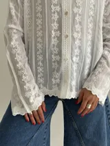 Camisa blanca de manga larga con delicados bordados florales verticales y detalles de encaje. Presenta cuello clásico, cierre frontal con botones y terminaciones festoneadas en los puños y el ruedo.