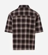 Camisa de corte boxy confeccionada en algodón, con diseño de cuadros escoceses y detalle de lettering bordado en el cuello. Presenta cuello camisero, mangas cortas y cierre frontal con botones.