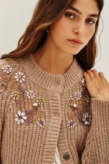 Cardigan de punto grueso en color azul, con cierre frontal de botones, mangas largas con puños de encaje blanco y detalle de bordado floral con pedrería brillante en el pecho.