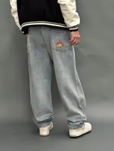 Pantalón de jean estilo baggy de corte ancho y relajado, color azul claro con lavado desgastado. Presenta un bordado en el bolsillo trasero derecho con un diseño de llamas y las siglas SDA.