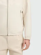 Chaqueta ligera con capucha, cierre frontal y puños elásticos, color beige claro. Presenta un diseño con costuras en forma de chevron en el pecho.