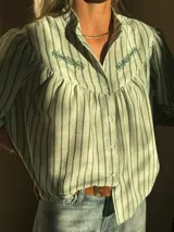 Camisa de corte holgado con diseño a rayas verticales en tonos verde claro y gris. Presenta cuello camisero, cierre frontal con botones y detalle de bordado floral en la parte superior del pecho. Las mangas son cortas y ligeramente abullonadas.