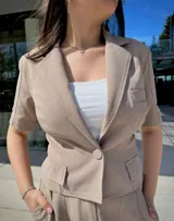 Blazer corto color beige, de manga corta, con solapas y cierre frontal con un botón. Presenta bolsillos de parche en la parte delantera.