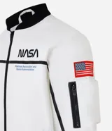 Campera tipo bomber blanca con detalles en negro, inspirada en uniformes espaciales de la NASA. Presenta parches con el logo de la NASA, la bandera de EE. UU. y texto en la manga que dice "Kennedy Space Center".