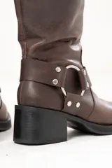 Bota biker de caña alta color marrón, confeccionada en cuero. Presenta hebilla en la parte superior, detalle de aro en la parte inferior y cierre lateral.