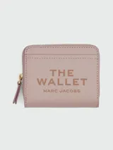 Billetera compacta con cierre de cremallera, color rosado pálido, con textura de cuero granulado. Presenta el texto "THE WALLET MARC JACOBS" estampado en el frente.