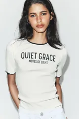 Camiseta de corte slim fit confeccionada en mezcla de algodón, con cuello redondo y manga corta. Presenta un diseño en color blanco con vivos negros en cuello y mangas, y un estampado frontal con el texto 'QUIET GRACE NOTES OF LIGHT'.