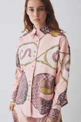 Camisa de corte recto y manga larga con cuello clásico y cierre frontal de botones. Presenta un estampado artístico de temática deportiva en tonos rosados, marrones y ocres, con un bolsillo frontal aplicado.