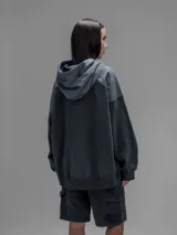 Hoodie oversize de color gris oscuro, confeccionado en frisa de algodón con recortes en textiles técnicos. Presenta capucha forrada, bolsillo frontal tipo canguro y un bolsillo adicional con solapa en la parte inferior.