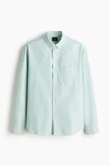 Camisa H&M de algodón Oxford con estampado de rayas verticales blancas y verdes, cuello americano, bolsillo frontal de parche y cierre central de botones.