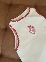 Chaleco sin mangas color blanco roto, con bordado de corazón anatómico en rojo en el pecho. Los bordes del cuello y las sisas tienen un ribete festoneado en color rojo.