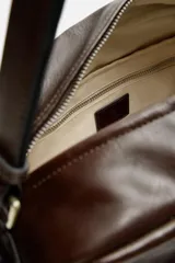 Bandolera marrón de piel con acabado envejecido, compartimento principal con cierre de cremallera, bolsillo interior plano y con cremallera, dos bolsillos frontales con botón a presión, dos bolsillos laterales con cremallera y asa de hombro ajustable.