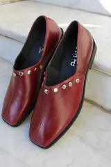 Zapato de cuero vacuno color bordó, con diseño de punta cuadrada y detalle de tachas metálicas planas en el borde superior. Presenta un taco geométrico bajo y costura central en el empeine.