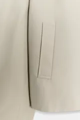 Trench corta color beige con cuello solapa, manga larga con trabilla y botón en los puños, bolsillos de vivo en el delantero y cierre frontal con botones oscuros.