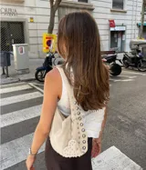 Bolso de hombro tipo hobo color negro, con acabado de gamuza o material similar. Decorado con tachas metálicas redondas plateadas a lo largo del borde inferior y en un dije colgante con forma de corazón.