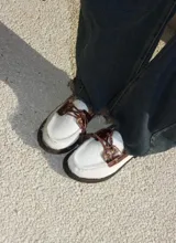 Mocasín de estilo náutico confeccionado en cuero, combinando una base de cuero con textura de pelo en color negro y detalles de cuero con textura croco en color marrón. Presenta cordones decorativos con borlas laterales y suela plana de goma.