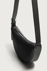 Bolso de hombro pequeño color negro con acabado liso, diseño semilunar y costura central. Cuenta con compartimento principal con cierre de cremallera, bolsillo interior con cierre y asa de hombro ajustable con hebilla.